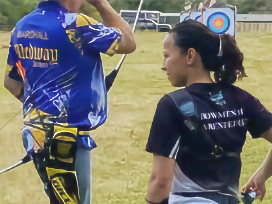 An archer in Medway uniform stands on the shooting line, and an archer in Darenteford uniform waits behin the shooting line, on a sunny weekend at the field.