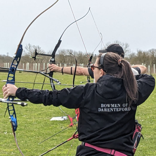 Archers are lined up with their bows raised, with the closest at full draw. Her jacket says 'Bowmen of Darenteford'.