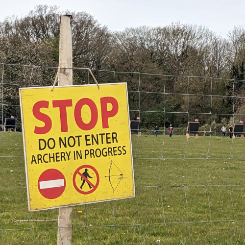 A safety sign is in the foreground, it says 'Stop, do not enter, archery in progress' and also has symbols below the writing. In the distance archers are approaching the bosses to collect their arrows.