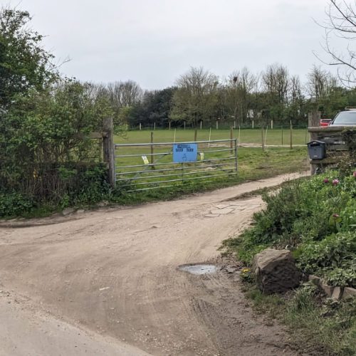 The entrance to the farm is on a rural dirt road. From the North, the entrance is on your right through a large metal gate. There's a blue sign on the gate that says 'Bluebell Barn Farm', and on the other side of the entrance there are flowers and a letterbox.