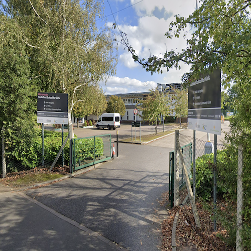 hedges and wire fences are on either side of the entrance. Black signs on each side read 'Welcome to Northfleet School for Girls.' Arrows point into the school grounds way for 'Main Reception, Car Park A, Northfleet Community Farm'. There is a speed limit of 5 miles per hour in the school grounds.