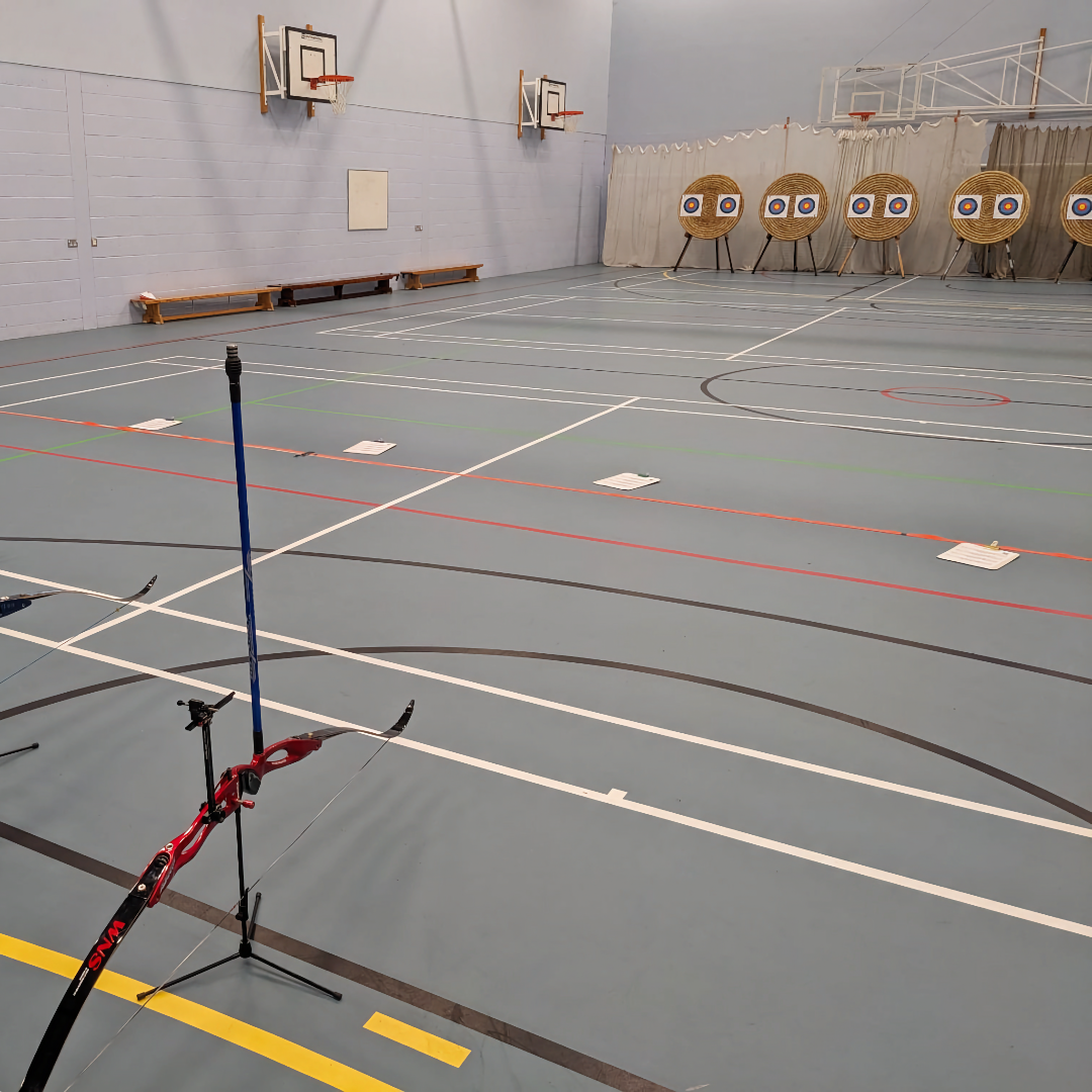 The shooting line is in the foreground, and a large curtain backstop hangs behind the bosses at the end of the hall. The floor is light grey with colourful line markings. There are scoresheets beside the orange shooting line, and benches against the walls.