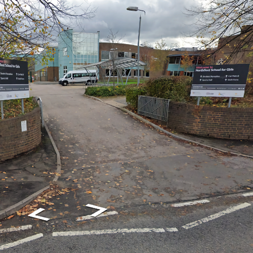 Curved low red brick walls are on either side of the entrance. A black sign reads 'Welcome to Northfleet School for Girls.' Arrows point the same way for 'Student Reception, Car Park B, Sports Hall, and Sixth Form'. There is a bike shelter and a school building along the driveway.